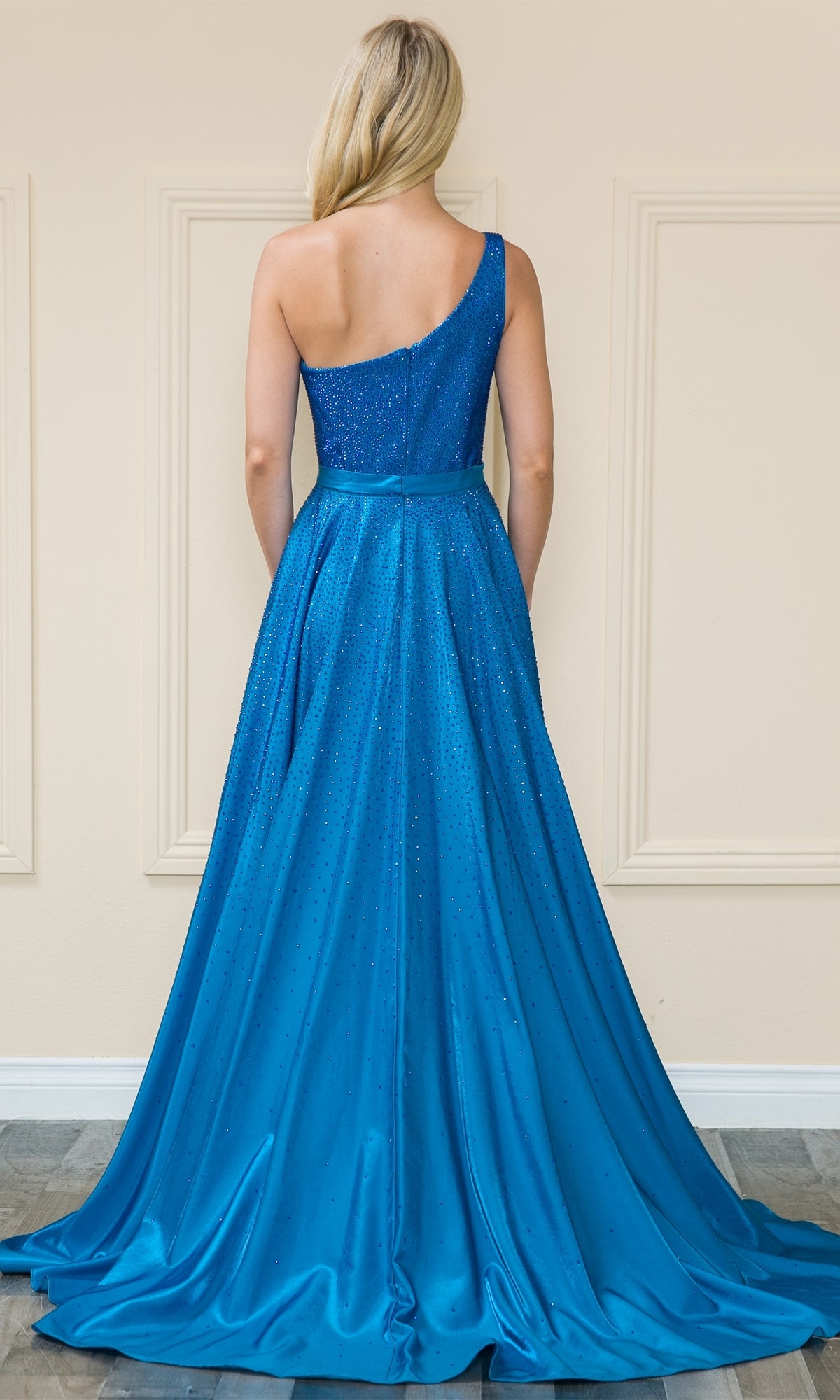 A woman with long blonde hair models the Poly USA 8914, a one-shoulder, floor-length A-line prom gown with beaded details and a fitted bodice, shown from the back.
